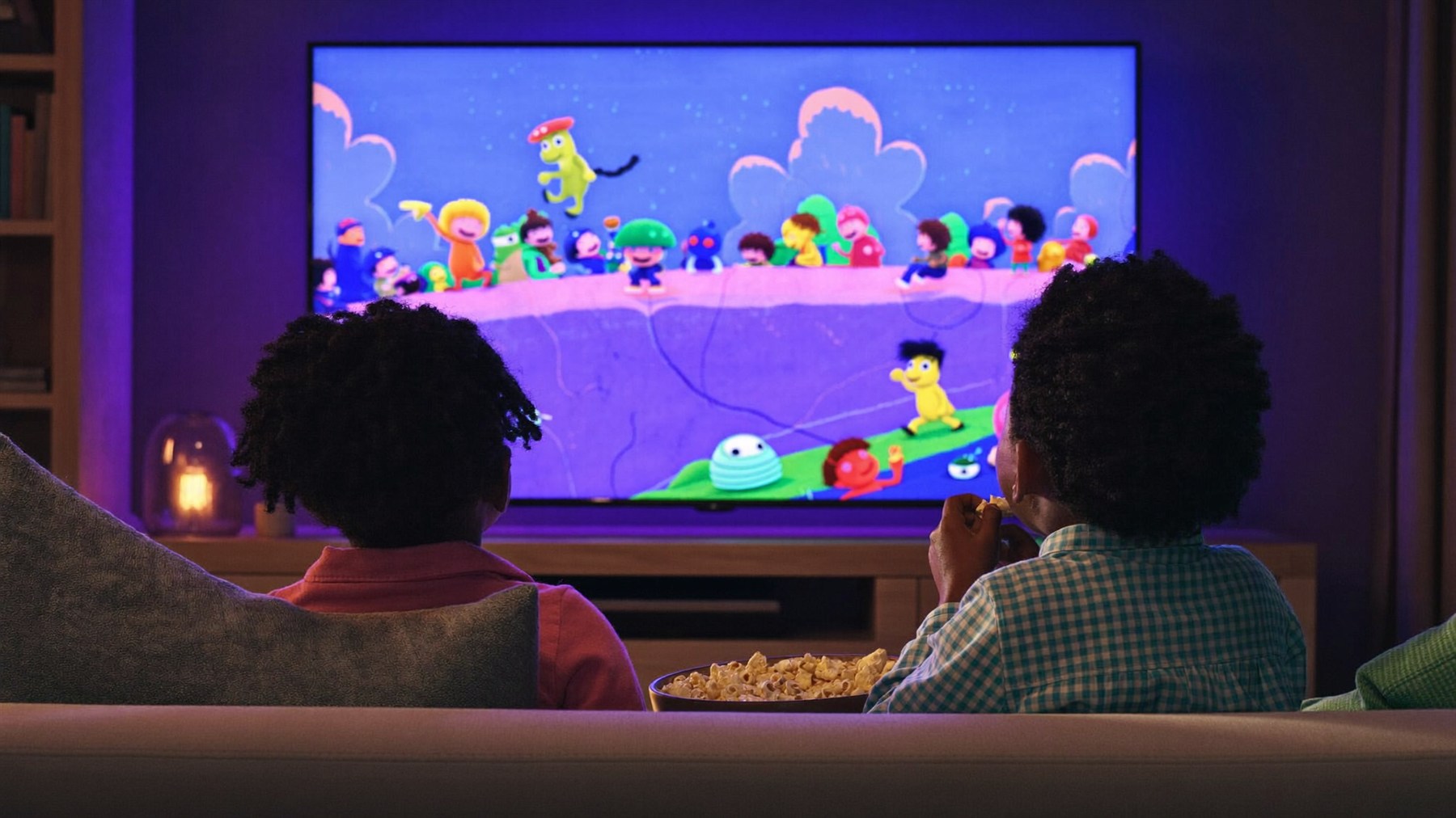 Two children seen from behind on a sofa with a bowl of popcorn, watching a colourful cartoon on the TV